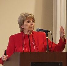 Charlene A. Caple speaking at a podium in a red blazer, microphone in hand, gesturing to the room.
